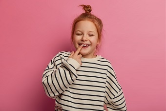 Joyful small redhaired female kid points at tooth, closes eyes and laughs happily, has bun knot, wears loose striped sweater, poses against rosy background, prepares for going to kindergarten